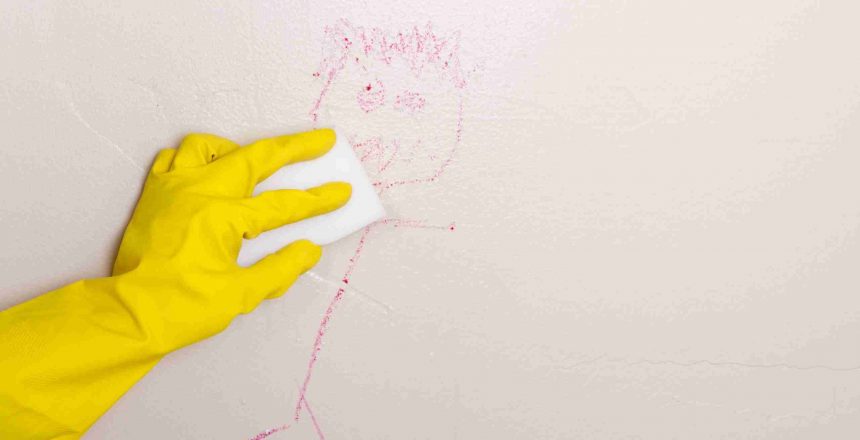 A hand with a yellow glove rubbing paraffin wax on the crayon drawing on a wall