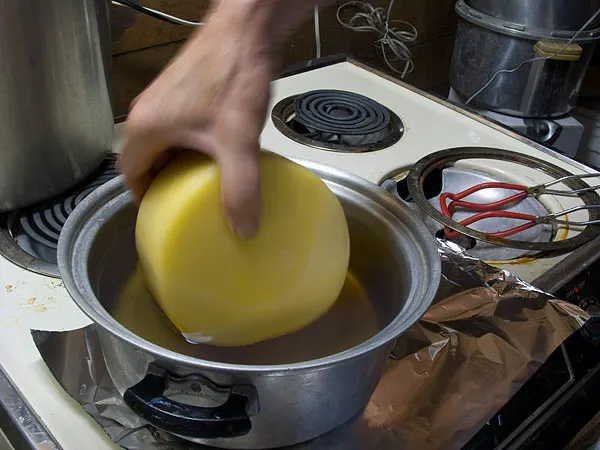 A person is dipping an entire wheel of cheese in a pot of melted paraffin wax