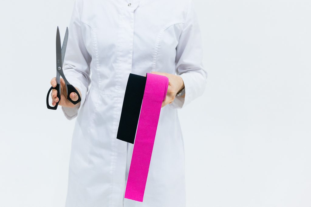 a physiotherapist wearing a white coat and holding black and pink athletic tape and scissors
