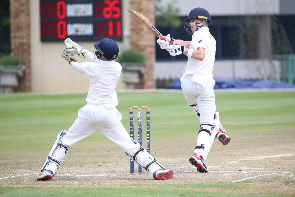 Two cricket players are playing cricket while in full gear, with the scoreboard in the background