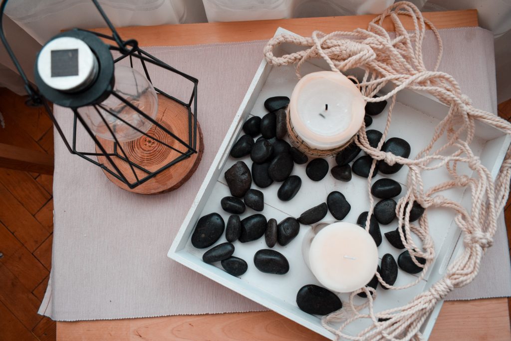 A pair of paraffin wax candles on a tray with black stones on a table right beside an oil lamp