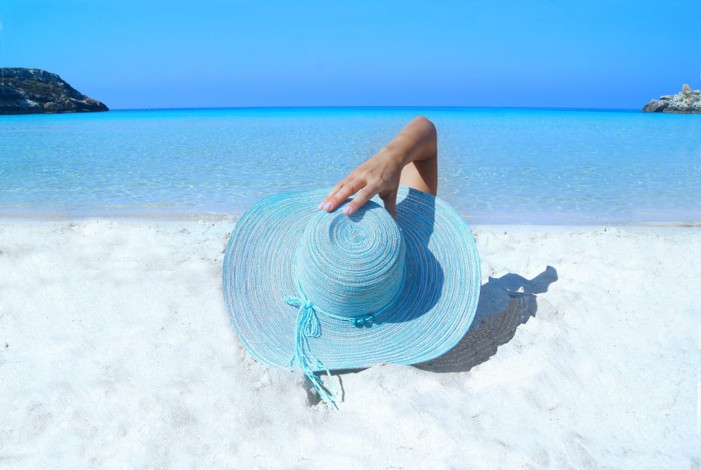 A person is enjoying the sun on a white beach and blue water while laying down and wearing a blue summer hat