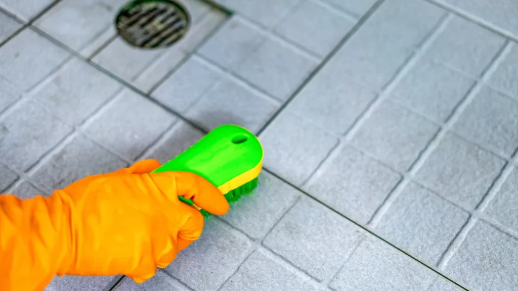 A person is scrubbing their tiles on the floor with some Epsom salt and a scrub brush while wearing an orange rubber glove