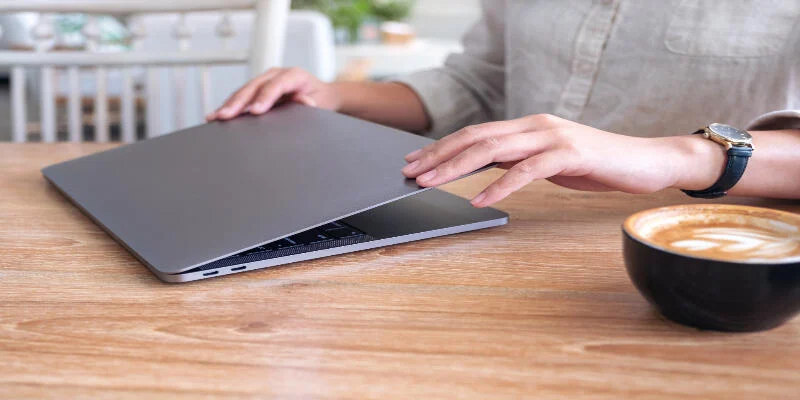 A physiotherapist is closing their laptop for the day on the wooden table while wearing a grey shirt and having a cup of coffee