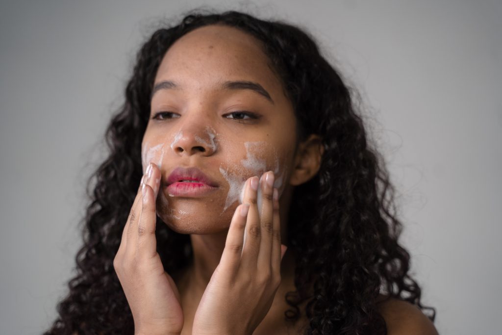 A young woman is massaging their face cleanser with some Epsom salt in front of a grey background
