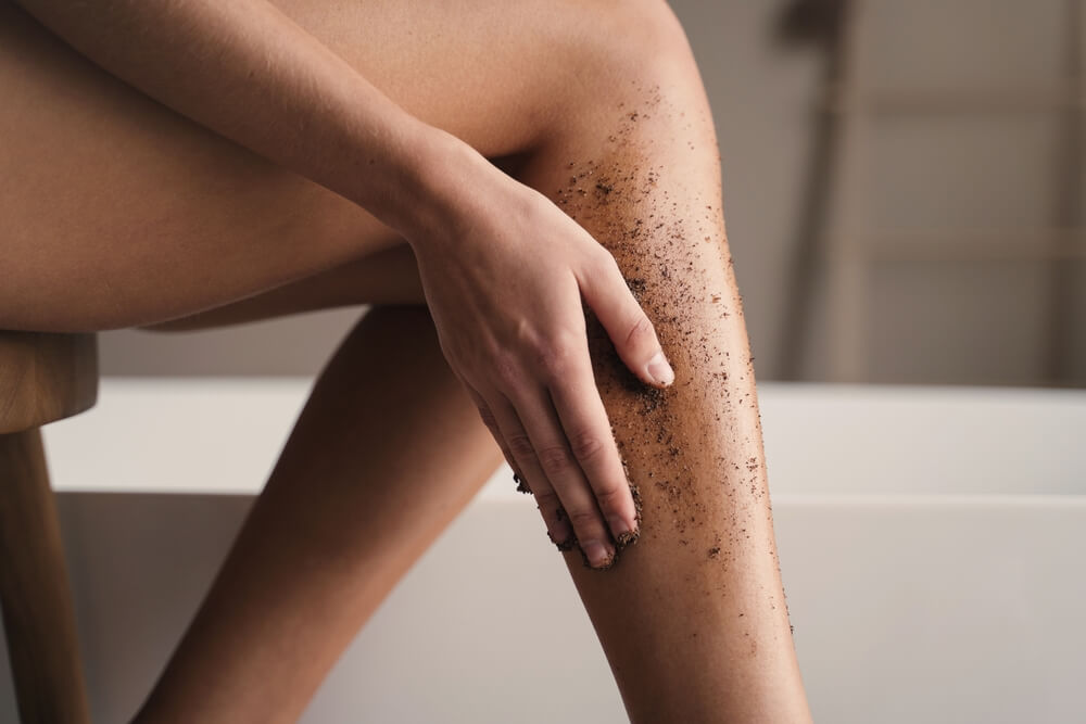 A person is scrubbing on an Epsom salt exfoliant on their legs by the side of their bathtub in their bathroom