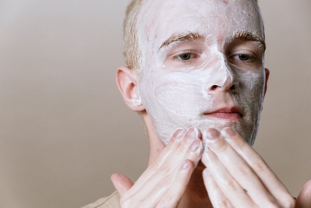 A man is putting on an Epsom salt face mask at home in front of a light brown background