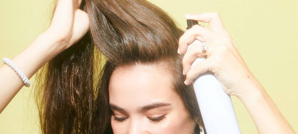 A woman is spraying hairspray all over her long hair in front of a yellow background