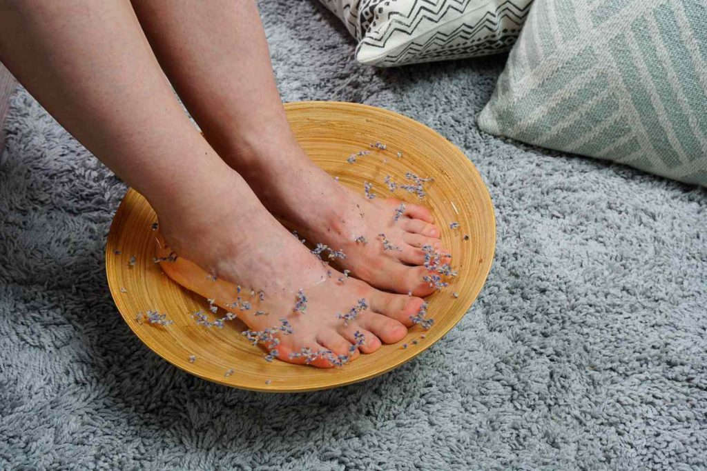 A lady is putting her feet in an Epsom salt foot bath while the shallow yellow bowl is on a plush blue shag with two pillows at its side