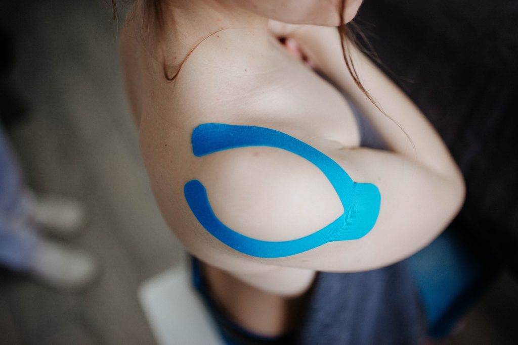 A person is showing the blue kinesio tape on their shoulder while holding their shirt over their shirt over their chest