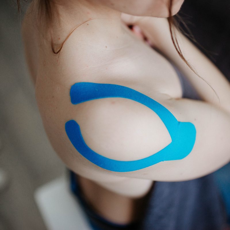 A person is showing the blue kinesio tape on their shoulder while holding their shirt over their shirt over their chest