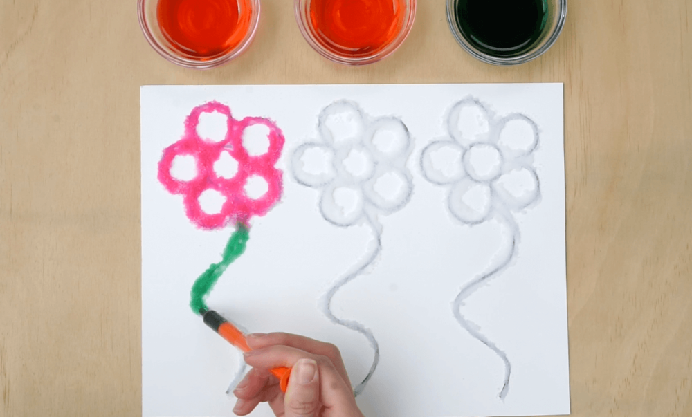 A person is making a painting of flowers using Epsom salt and coloured water on paper on a wooden table