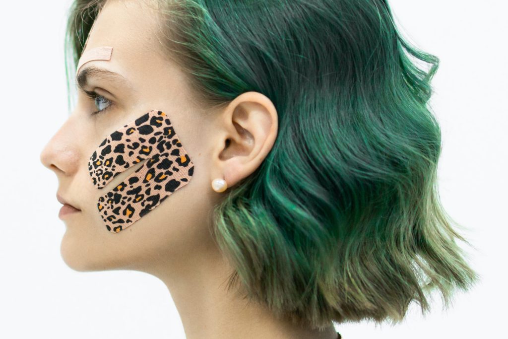 A lady with short green hair has KT tape in a leopard print on their cheek, her face in view while in front of a white background