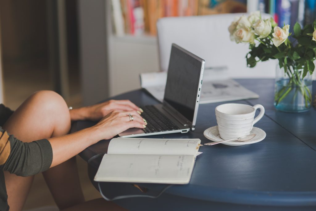 An employee of a physiotherapy clinic checking the policies of possible suppliers while sitting at a blue table, with an open laptop, notebook, catalogue, cup of coffee, and a vase full of roses on it