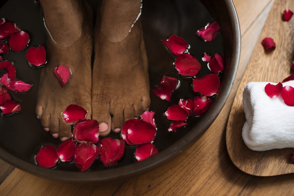 Image of a person having themselves an Epsom salt foot bath in a wooden bowl on a wooden flooring, with rose petals in the water and a towel ready at the side
