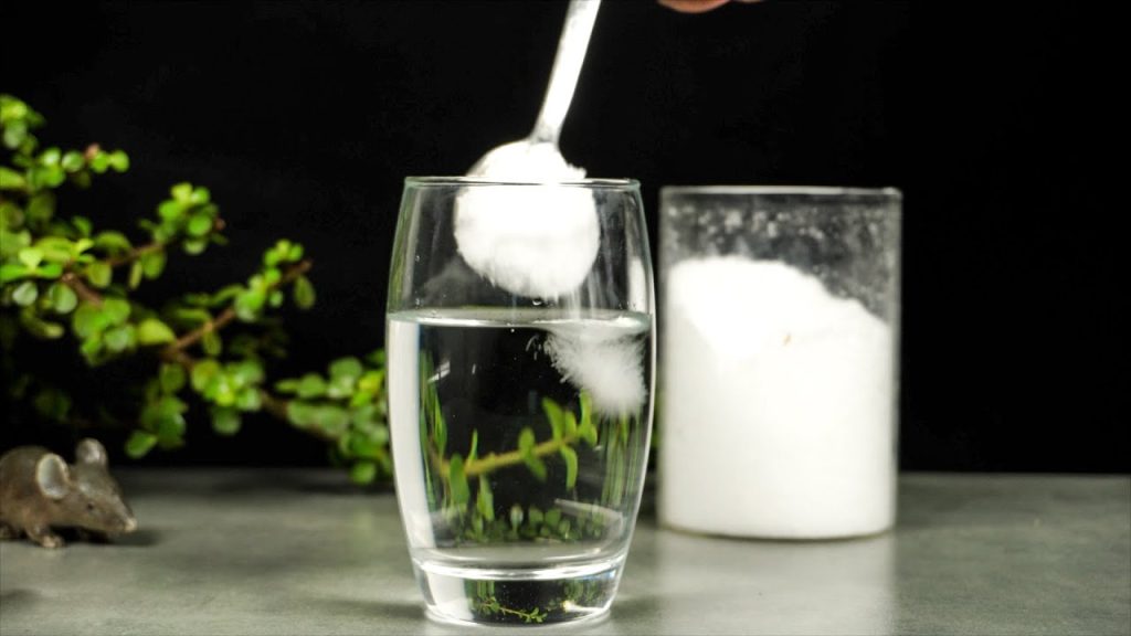 A person is mixing in a spoonful of Epsom salt in a glass of water on a wooden table while there's a canister of Epsom salt and a plant in the background