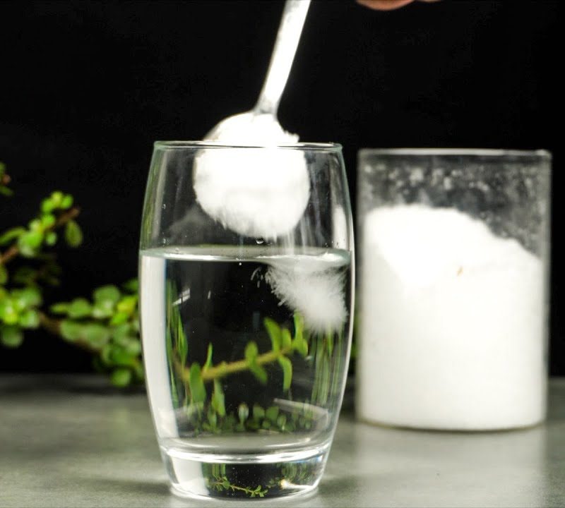 A person is mixing in a spoonful of Epsom salt in a glass of water on a wooden table while there's a canister of Epsom salt and a plant in the background