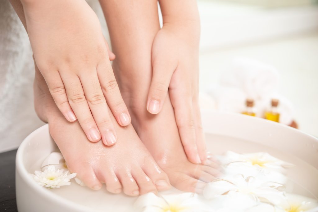 A woman is submerging their feet in a ceramic bowl for their Epsom salt foot bath with flowers in the water and bottles of oils in the background