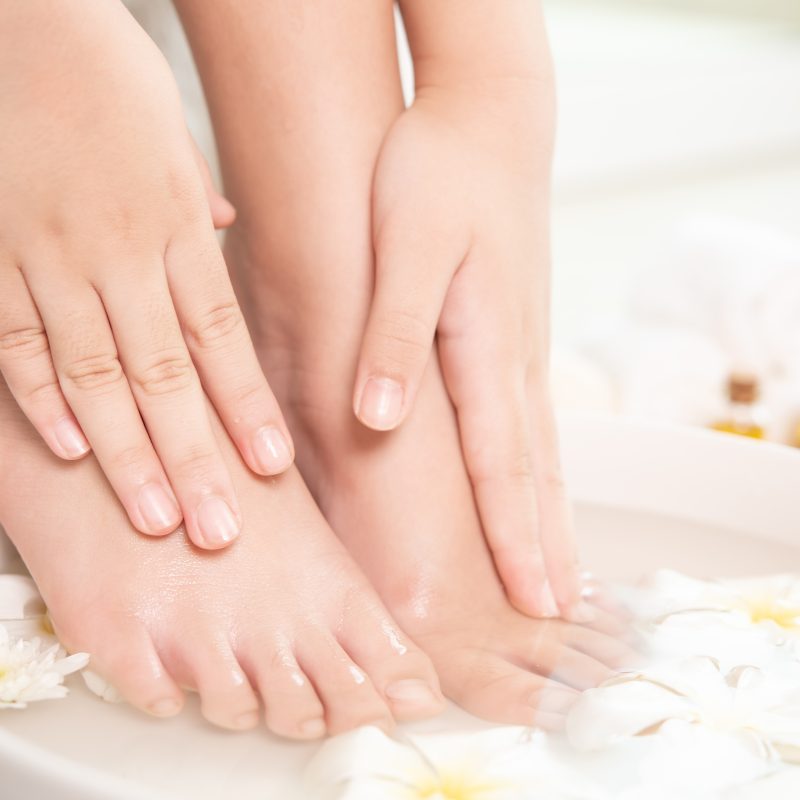 A woman is submerging their feet in a ceramic bowl for their Epsom salt foot bath with flowers in the water and bottles of oils in the background