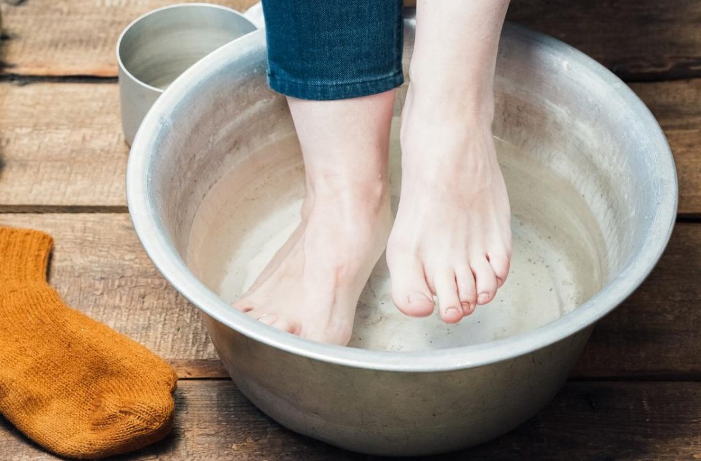 A person is trying out a simple Epsom salt foot bath in a metal basin on wooden flooring while a small water basin is set aside and their sock ready by their side