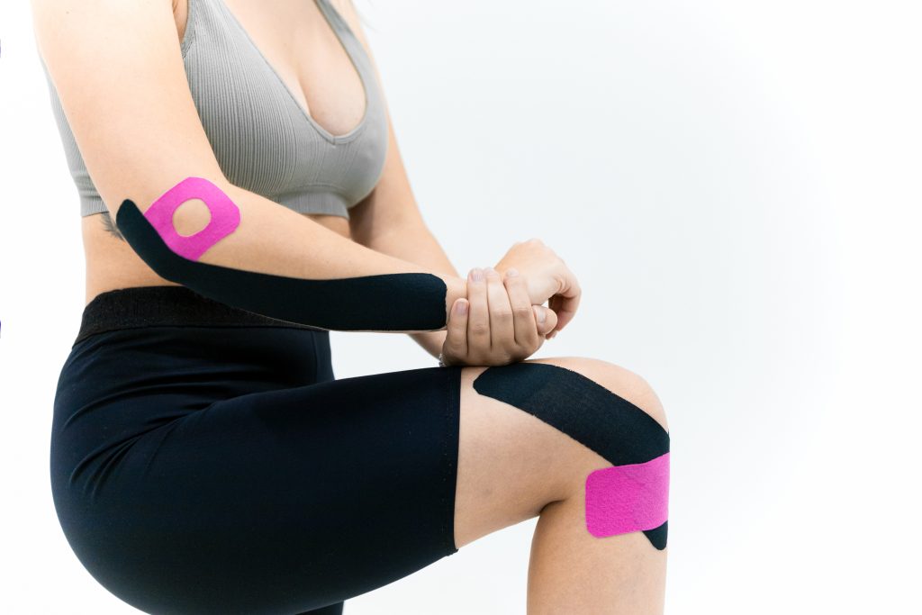 A woman has both pink and black KT tape on her knee and forearm, her leg propped up while her arm is on it for support