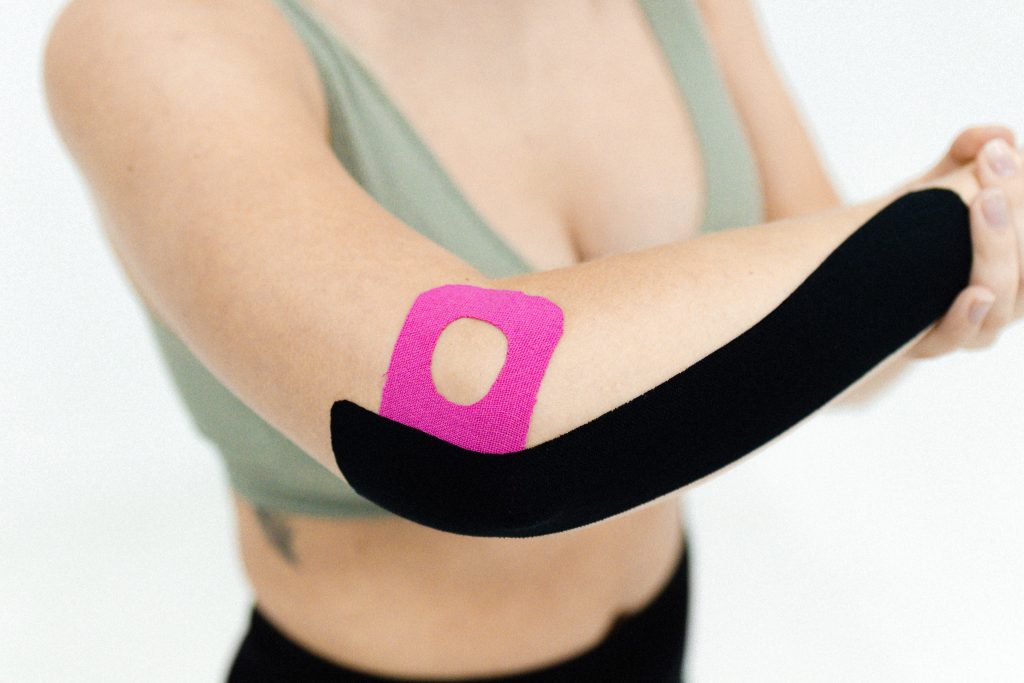A lady has black and pink kinesio tape on her forearm and elbow, while in front of a white background and wearing a grey top and black shorts