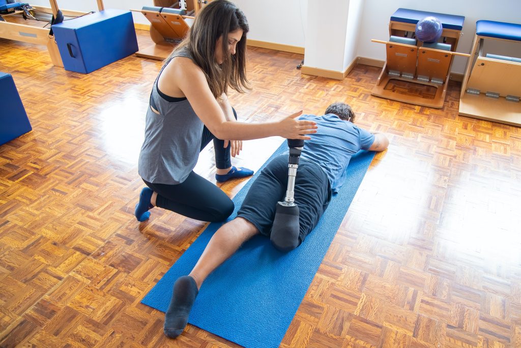 A physiotherapist is holding their client's attached prosthetic leg up while the client is laying down on their stomach on a blue mat on the floor