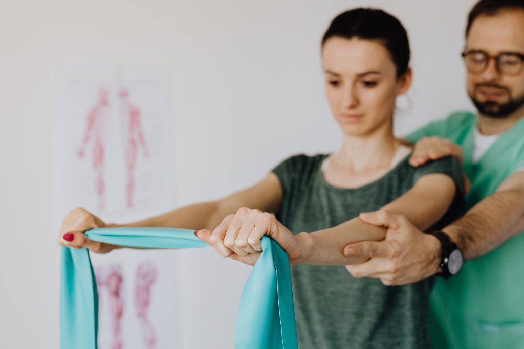 A physiotherapist is with a client, helping them stretch their arms with a band