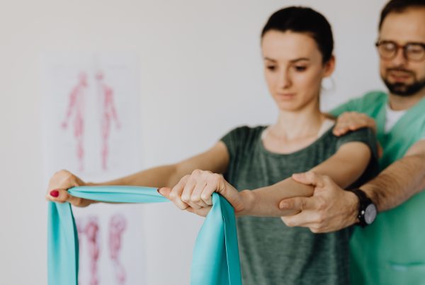 A physiotherapist is with a client, helping them stretch their arms with a band