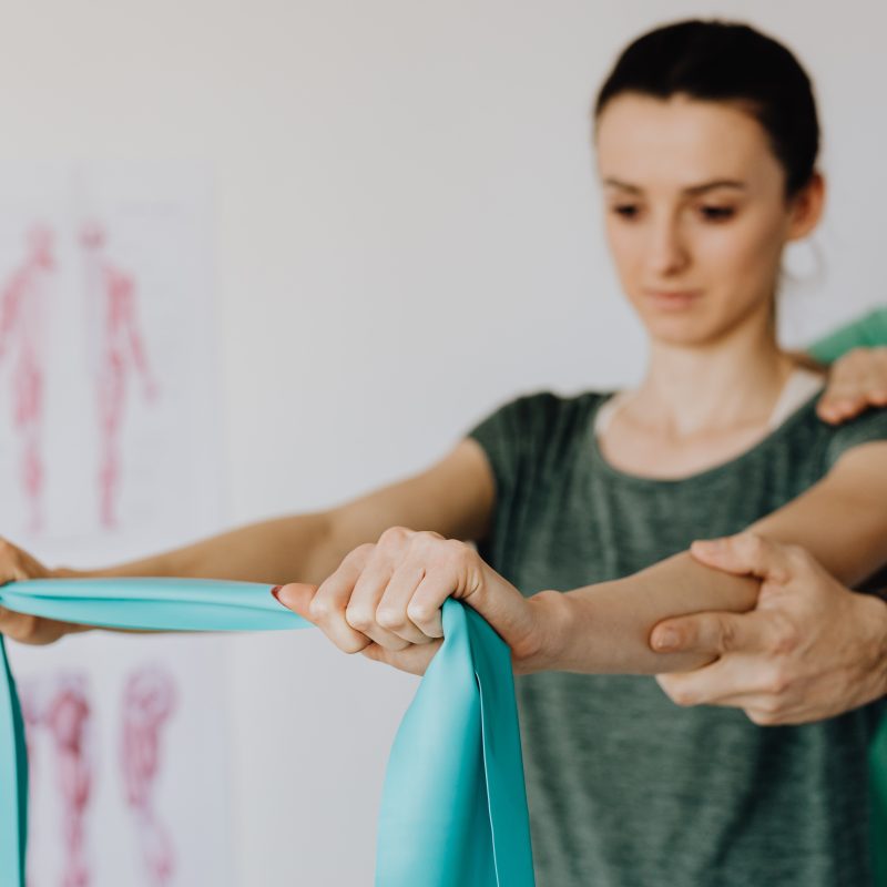 A physiotherapist is with a client, helping them stretch their arms with a band