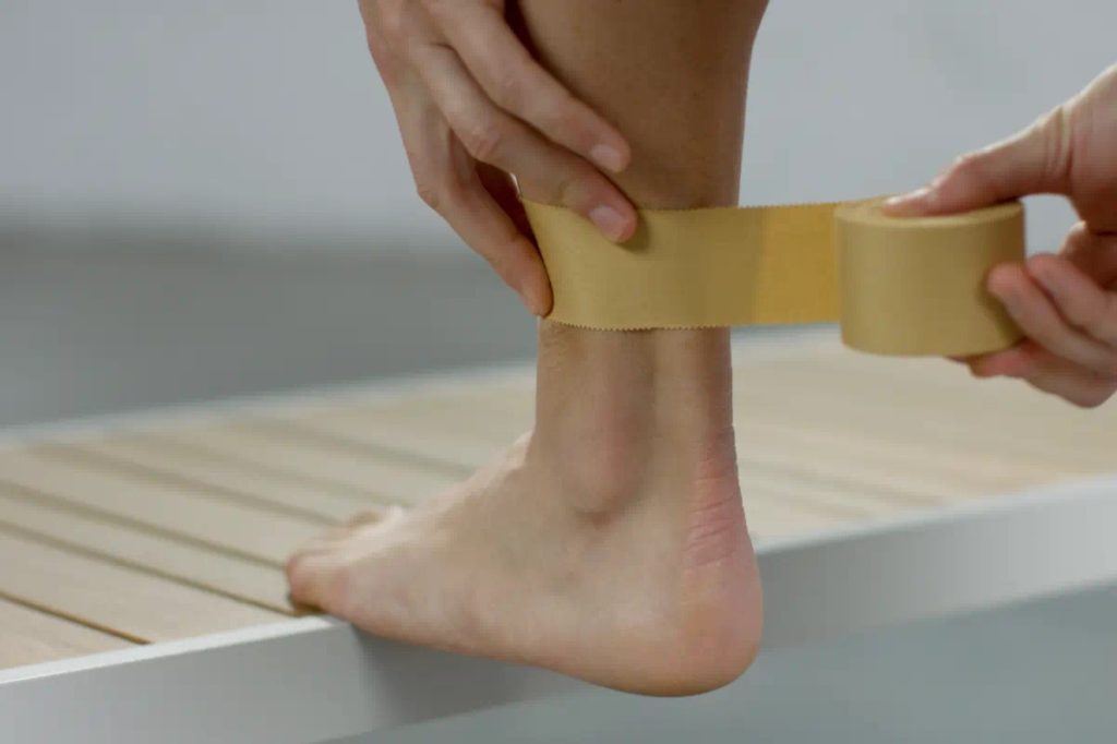 A person has their foot on top of a wooden brown and white bench while they strap on some rigid tape above their ankle
