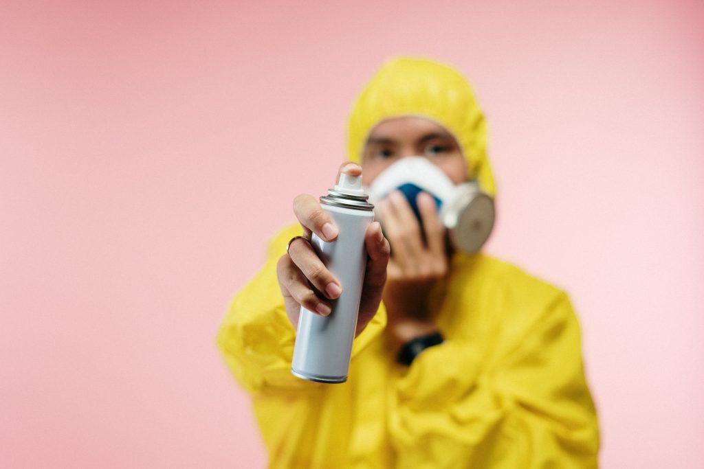 A person is wearing a gas mask and a hazmat suit while handling a can of formaldehyde in front of a pink background
