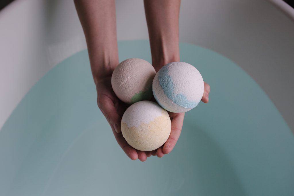 A person is holding out three bath bombs in different colours over a bathtub full of water