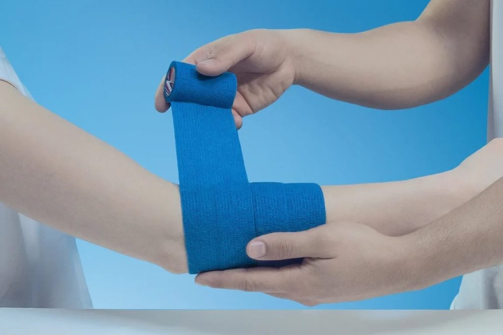 A person is wrapping the arm of another person with blue cohesive bandages while on top of a table and in front of a light blue background