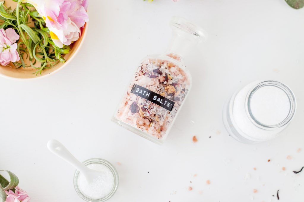 A jar of bath salts with a black label is on its side on a white table while being surrounded by flowers, other glasses, and its other ingredients