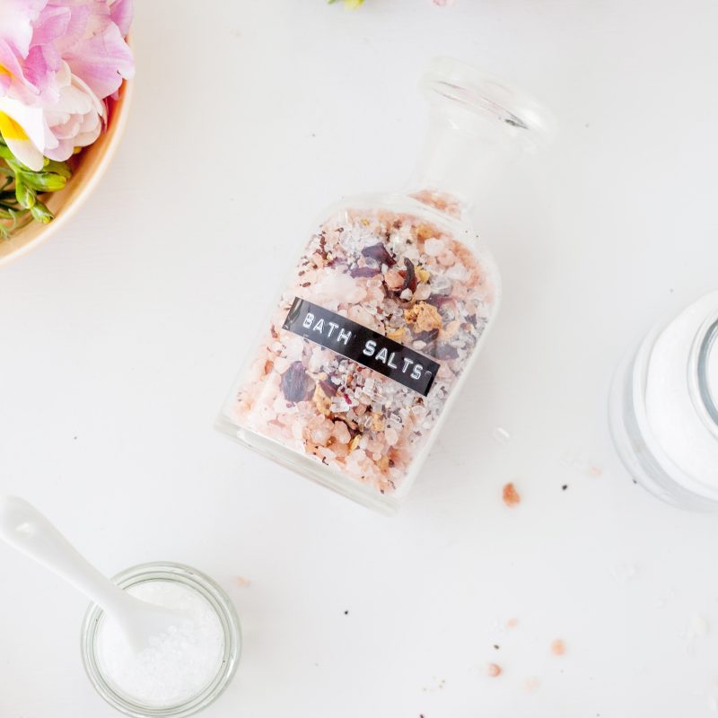 A jar of bath salts with a black label is on its side on a white table while being surrounded by flowers, other glasses, and its other ingredients