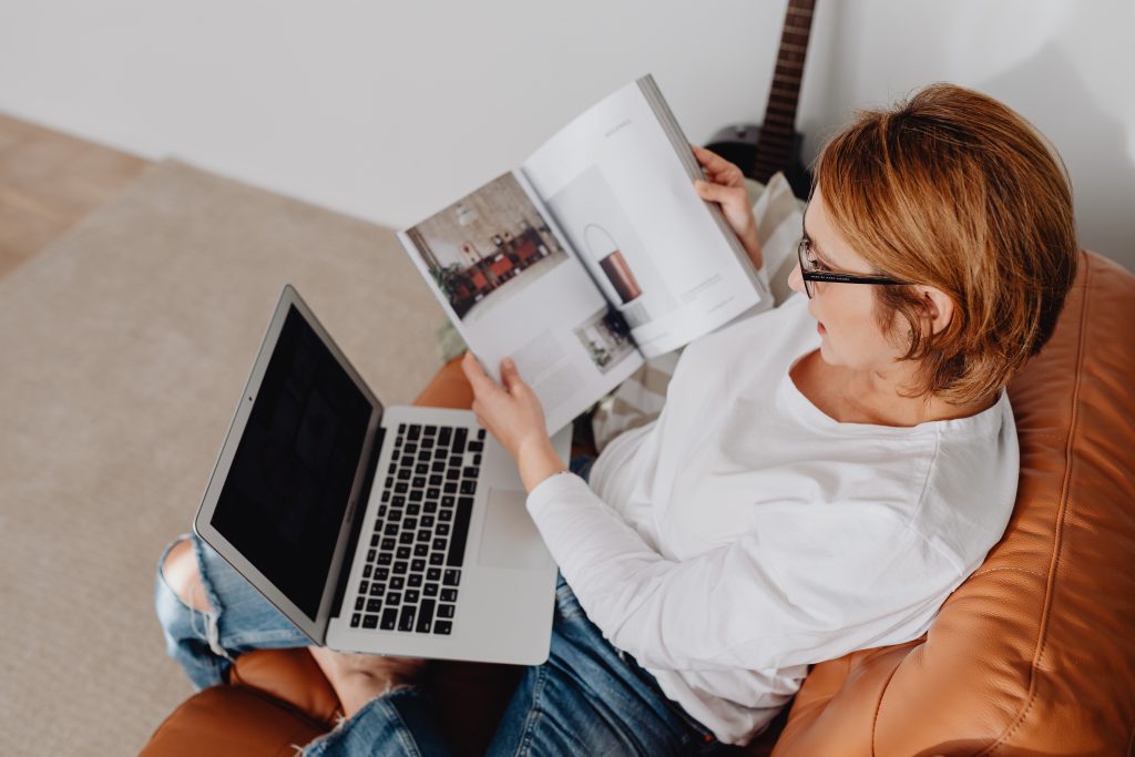 A woman is verifying the items on a printed catalogue with the supplier's website while sitting down on a comfy chair and wearing glasses