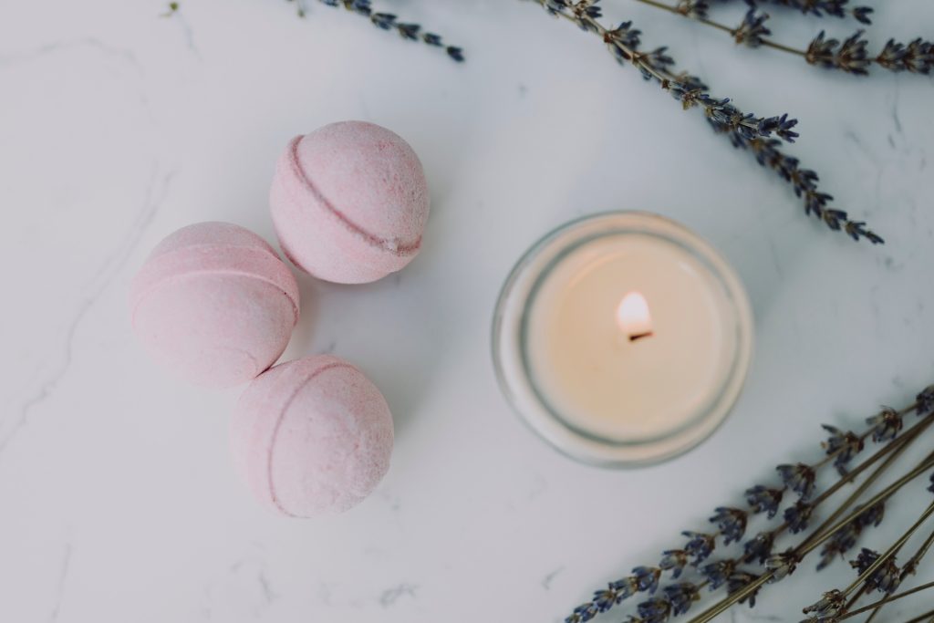 Three pink bath bombs, sprigs of herbs, and a lit candle in a jar are all on top of a white marble table