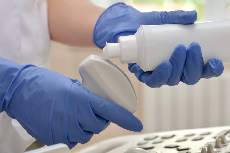 A sonographer is putting ultrasound gel on the transducer while wearing disposable blue gloves and a white coat