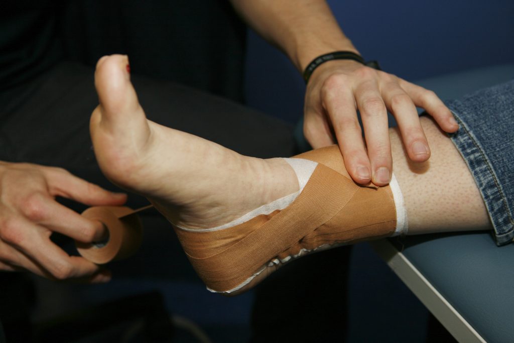 A medical professional is wrapping the ankle of a jeans-wearing client while the client has their foot raised on a physiotherapy bed