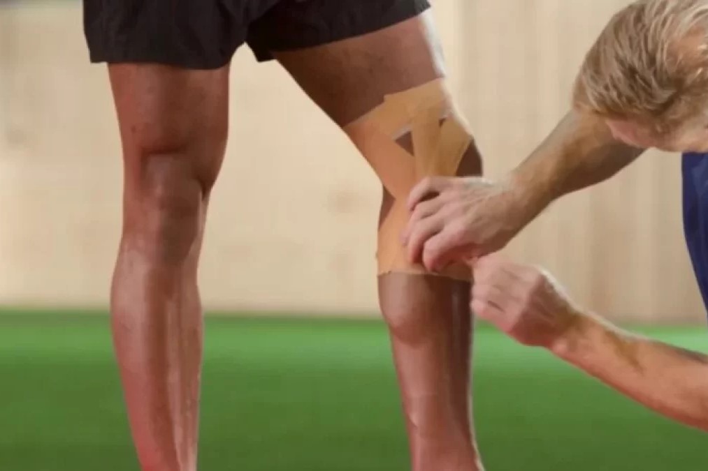 A medical professional is taping rigid tape on an athlete's knee and upper calf while the athlete is wearing black shorts and standing in the middle of the field