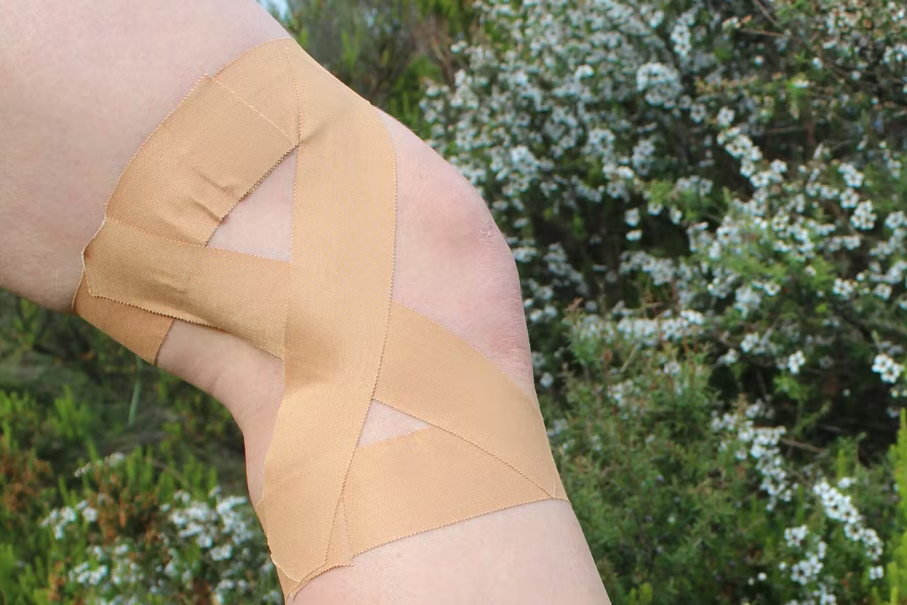 A person is showing the rigid tape on and around their knee while in front of some leaved and white flowers