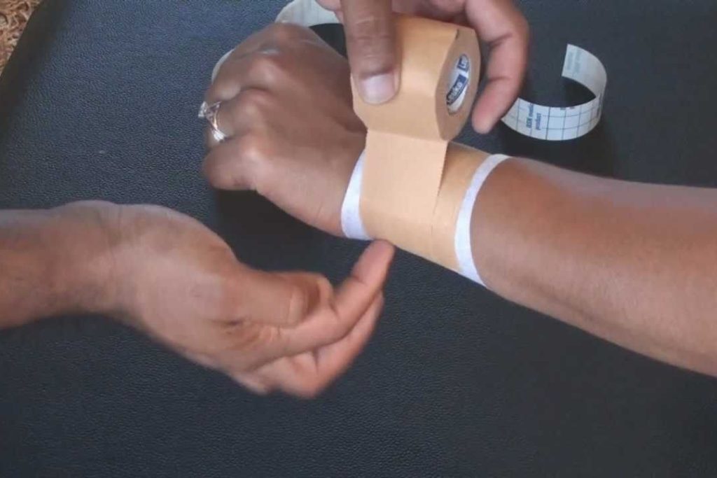 A client has their hand on top of a dark blue or black table so that their hand can get wrapped with rigid tape by a physiotherapist