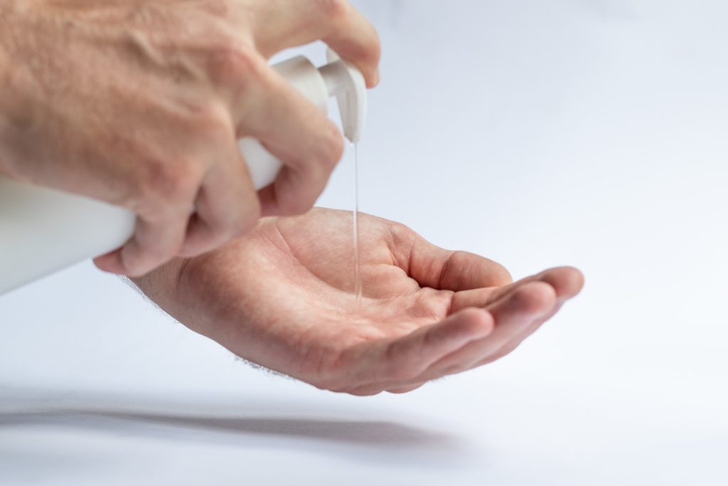 One hand is using a bottle with a pump cap to dispense disinfectant/antiseptic in the other hand while on a white table and in front of a white background