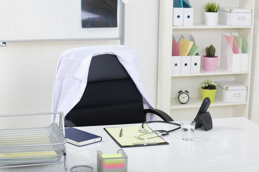 A physiotherapist's desk is in a white room with a whiteboard and book case in the background, and their stuff scattered around the desk and shelves like a notebook, phone, white coat, clock, plants, and file folders