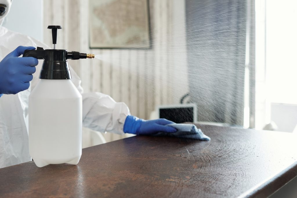 A person in blue gloves and white protective gear is spraying a table with a disinfectant and wiping it down with a cloth after