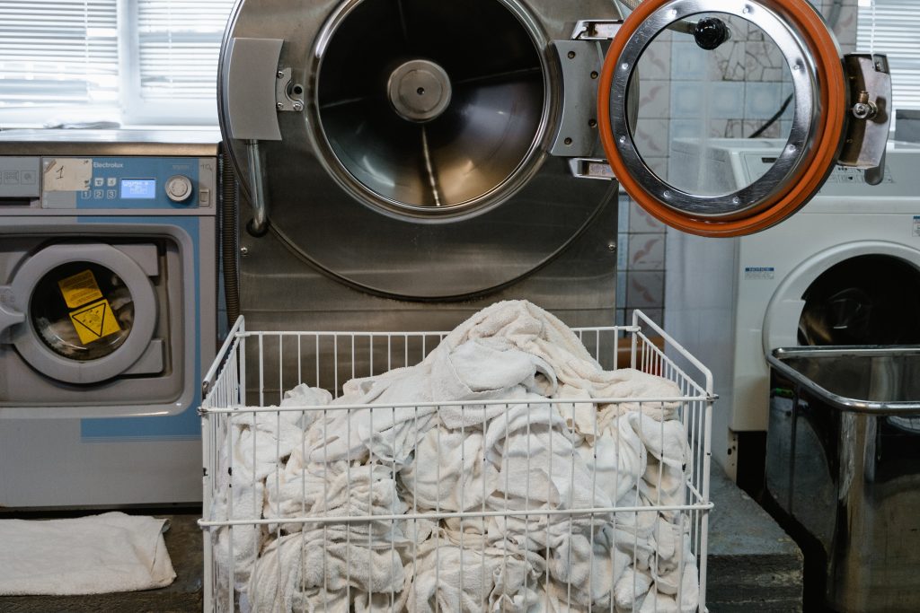 A huge washing machine in between smaller machines is being loaded with a quats-based disinfectant and a cart full of dirty white linens