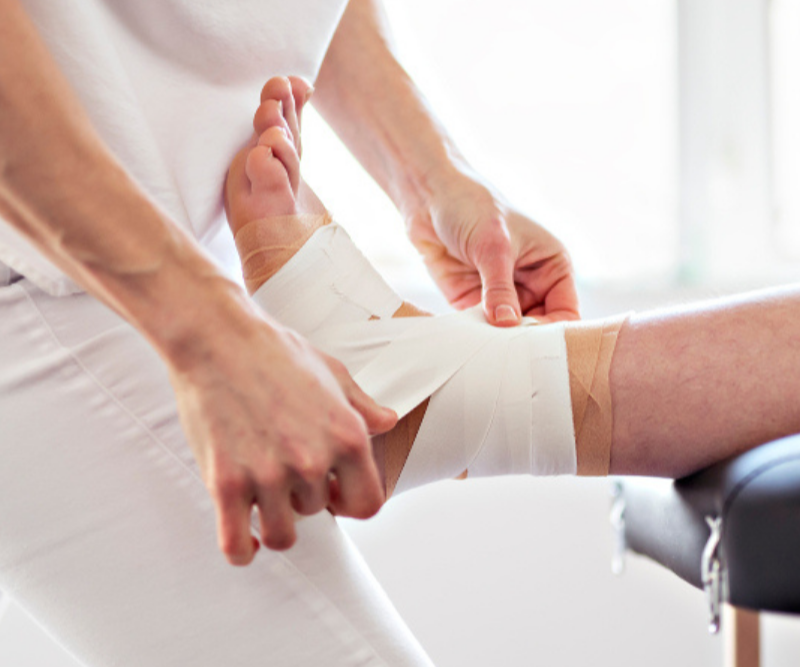 A physiotherapist is strapping rigid tape on a client's ankle while wearing a white shirt and white pants and the client's leg is on the edge of an examination table