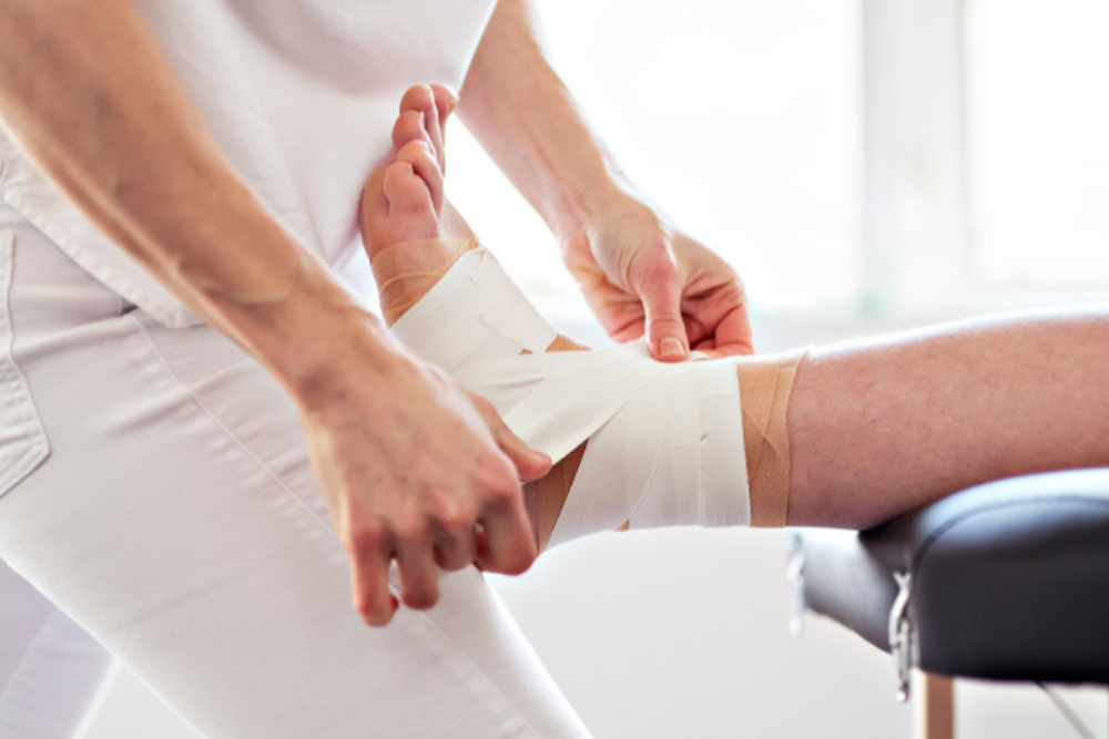 A physiotherapist is strapping rigid tape on a client's ankle while wearing a white shirt and white pants and the client's leg is on the edge of an examination table