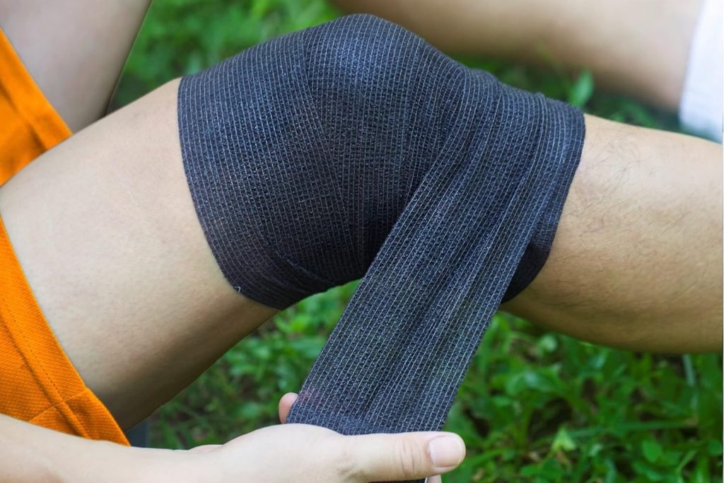 A person is sitting down on the grass and wearing bright orange shorts while bandaging their knee with black cohesive bandage